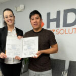 A woman wearing glasses, a white blouse, and a black blazer, and a man in a dark shirt smile while posing in an office at HDS Biz Solutions. Both hold certificates or official documents in front of the company’s gray and orange logo on the wall behind them. The setting is an office with light-colored walls, a paneled ceiling, and chairs in the background.