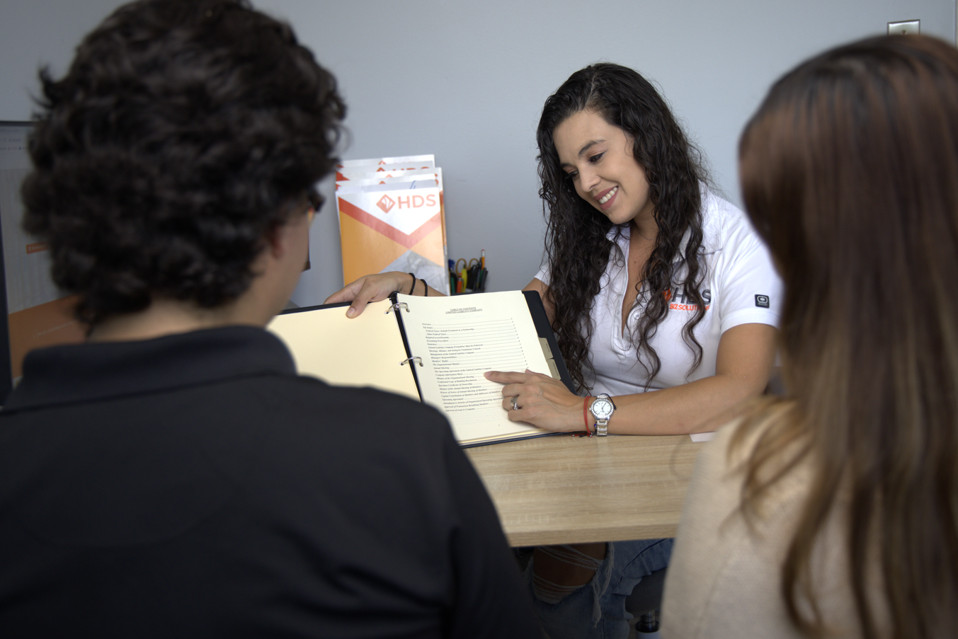 A professional advisor from HDS Biz Solutions, wearing a white shirt with the company logo, smiles while presenting and explaining a document in a black folder to a young couple seated across the table. The couple listens attentively, with the woman slightly leaning in. In the background, HDS promotional materials are visible, reinforcing the professional and supportive environment of the consultation. The scene reflects a welcoming and informative setting for business and licensing services.