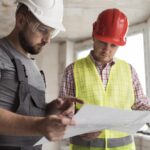 contratistas con chalecos reflectantes y cascos de seguridad revisan un proyecto en una tableta en el interior de un edificio en construcción; una de las personas sostiene planos