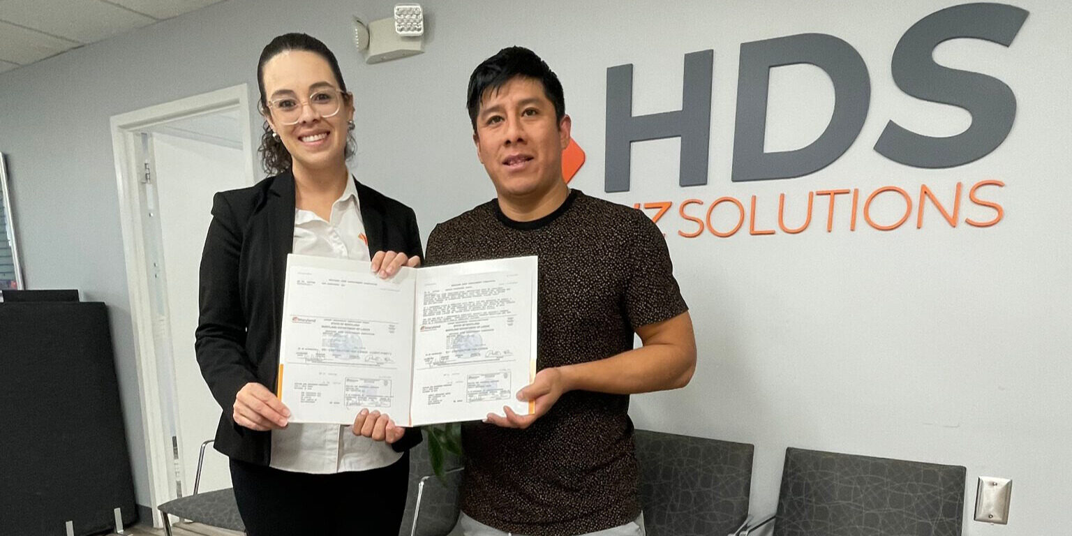 A woman wearing glasses, a white blouse, and a black blazer, and a man in a dark shirt smile while posing in an office at HDS Biz Solutions. Both hold certificates or official documents in front of the company’s gray and orange logo on the wall behind them. The setting is an office with light-colored walls, a paneled ceiling, and chairs in the background.