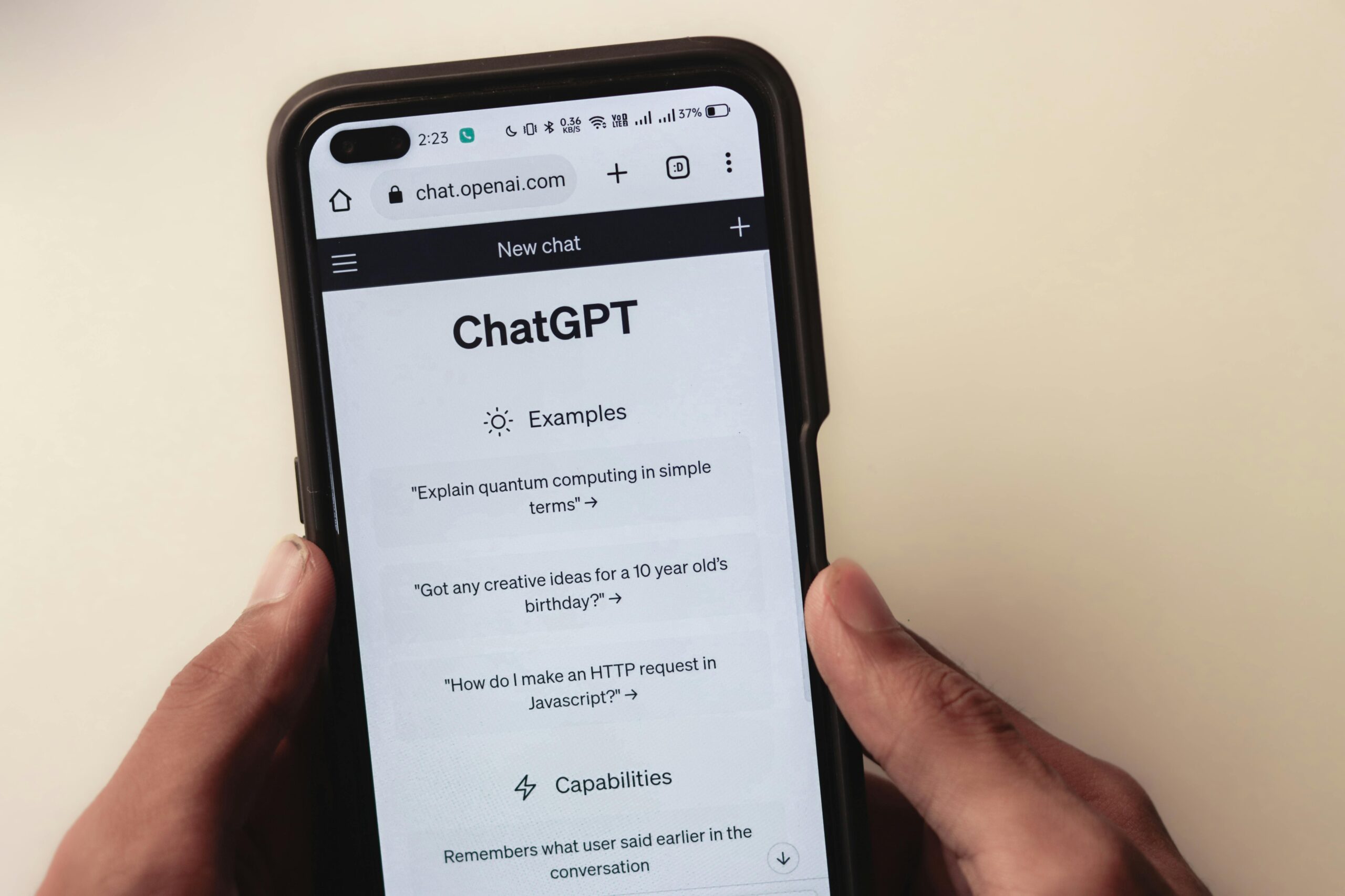 Persona sosteniendo un teléfono móvil que muestra la página de inicio de ChatGPT en el sitio web chat.openai.com, con ejemplos de preguntas visibles en la pantalla
