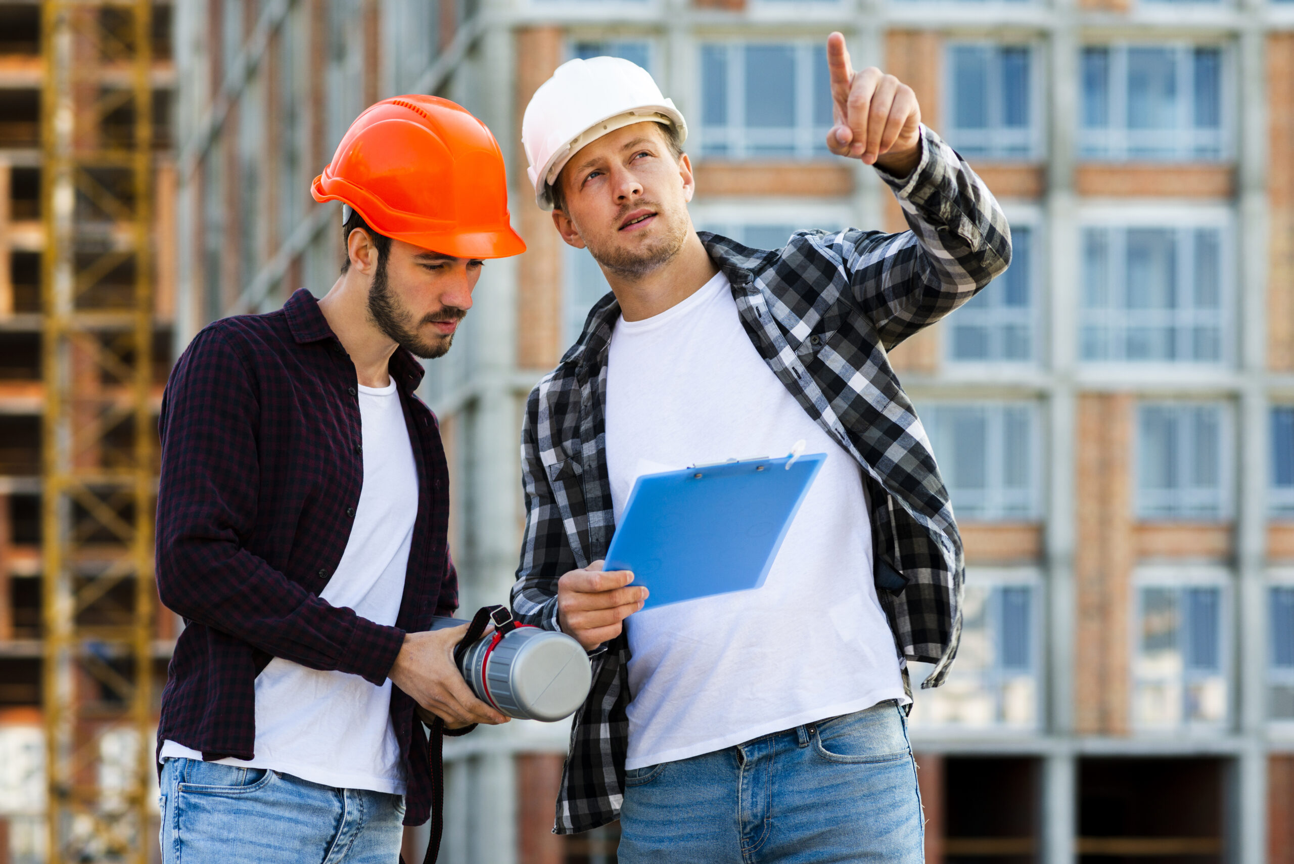 Hombre con casco blanco y chaleco reflectante naranja trabajando en una obra en construcción, revisando planos sobre una mesa mientras usa una computadora portátil. Al fondo, dos trabajadores más con cascos y chalecos naranjas conversan y se mueven por el espacio