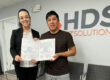 A woman wearing glasses, a white blouse, and a black blazer, and a man in a dark shirt smile while posing in an office at HDS Biz Solutions. Both hold certificates or official documents in front of the company’s gray and orange logo on the wall behind them. The setting is an office with light-colored walls, a paneled ceiling, and chairs in the background.