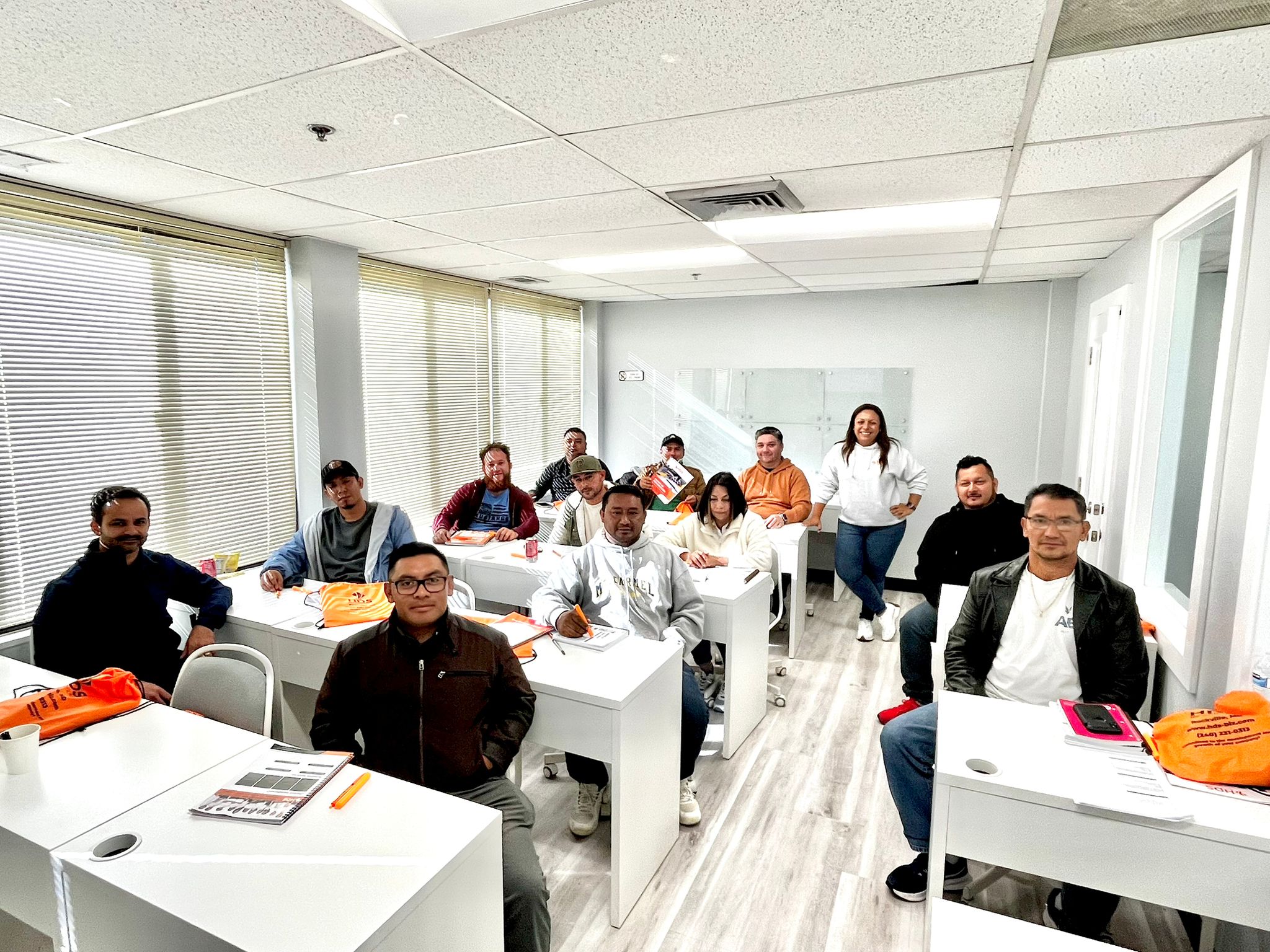 Grupo de contratistas latinos participando en una clase presencial de preparación para la licencia de contratista en Virginia, en un aula moderna y bien iluminada. La instructora sonríe de pie al fondo, mientras los estudiantes están atentos con materiales de estudio y carpetas naranjas de HDS Biz Solutions sobre sus escritorios