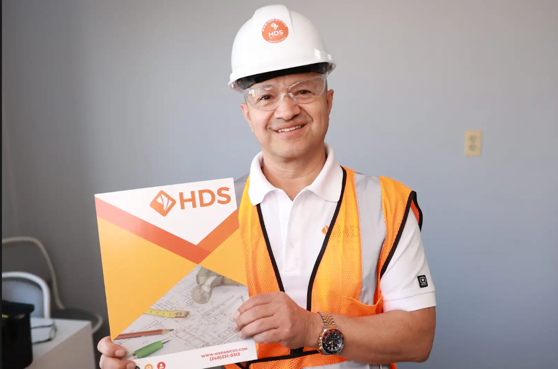 Un hombre sonriente con casco blanco y chaleco de seguridad naranja sostiene una carpeta con el logo de HDS. La carpeta muestra planos de construcción y herramientas, destacando un entorno profesional relacionado con la industria de la construcción, licencia de contratista o contractor license