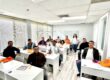 A group of Latino contractors attending an in-person contractor license prep class in Virginia, inside a modern and well-lit classroom. The instructor smiles while standing at the back, as the students stay focused with study materials and orange HDS Biz Solutions folders on their desks