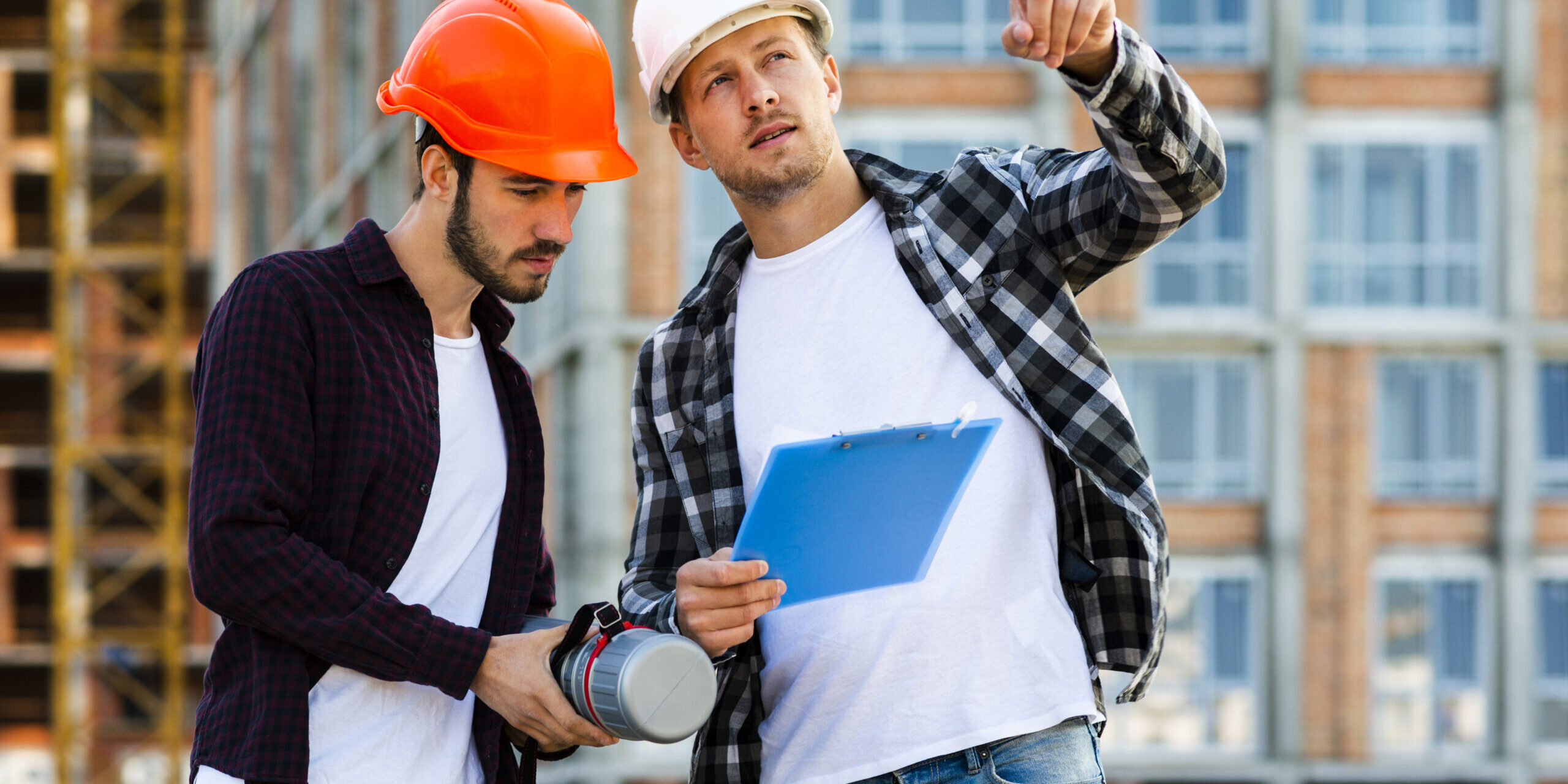 Hombre con casco blanco y chaleco reflectante naranja trabajando en una obra en construcción, revisando planos sobre una mesa mientras usa una computadora portátil. Al fondo, dos trabajadores más con cascos y chalecos naranjas conversan y se mueven por el espacio