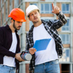 Hombre con casco blanco y chaleco reflectante naranja trabajando en una obra en construcción, revisando planos sobre una mesa mientras usa una computadora portátil. Al fondo, dos trabajadores más con cascos y chalecos naranjas conversan y se mueven por el espacio