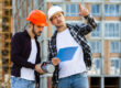 Hombre con casco blanco y chaleco reflectante naranja trabajando en una obra en construcción, revisando planos sobre una mesa mientras usa una computadora portátil. Al fondo, dos trabajadores más con cascos y chalecos naranjas conversan y se mueven por el espacio
