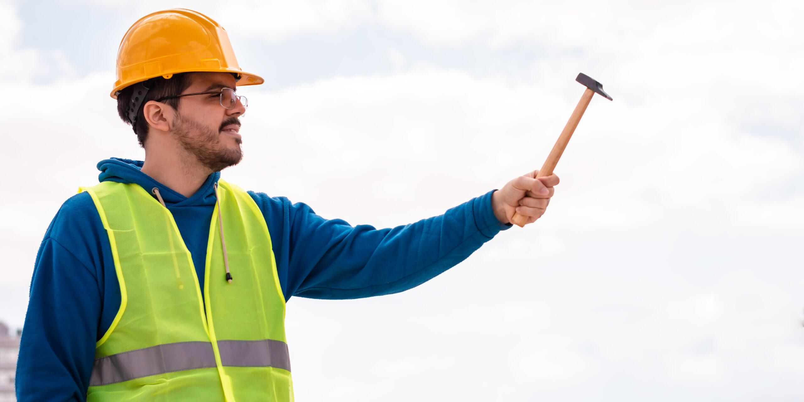Trabajador de construcción con chaleco de seguridad y casco, sosteniendo un martillo al aire libre, representando profesionalismo y preparación en el sitio de trabajo