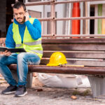 Construction worker in a yellow safety vest sitting on a bench, talking on the phone and reviewing a notebook, with a yellow hard hat beside him