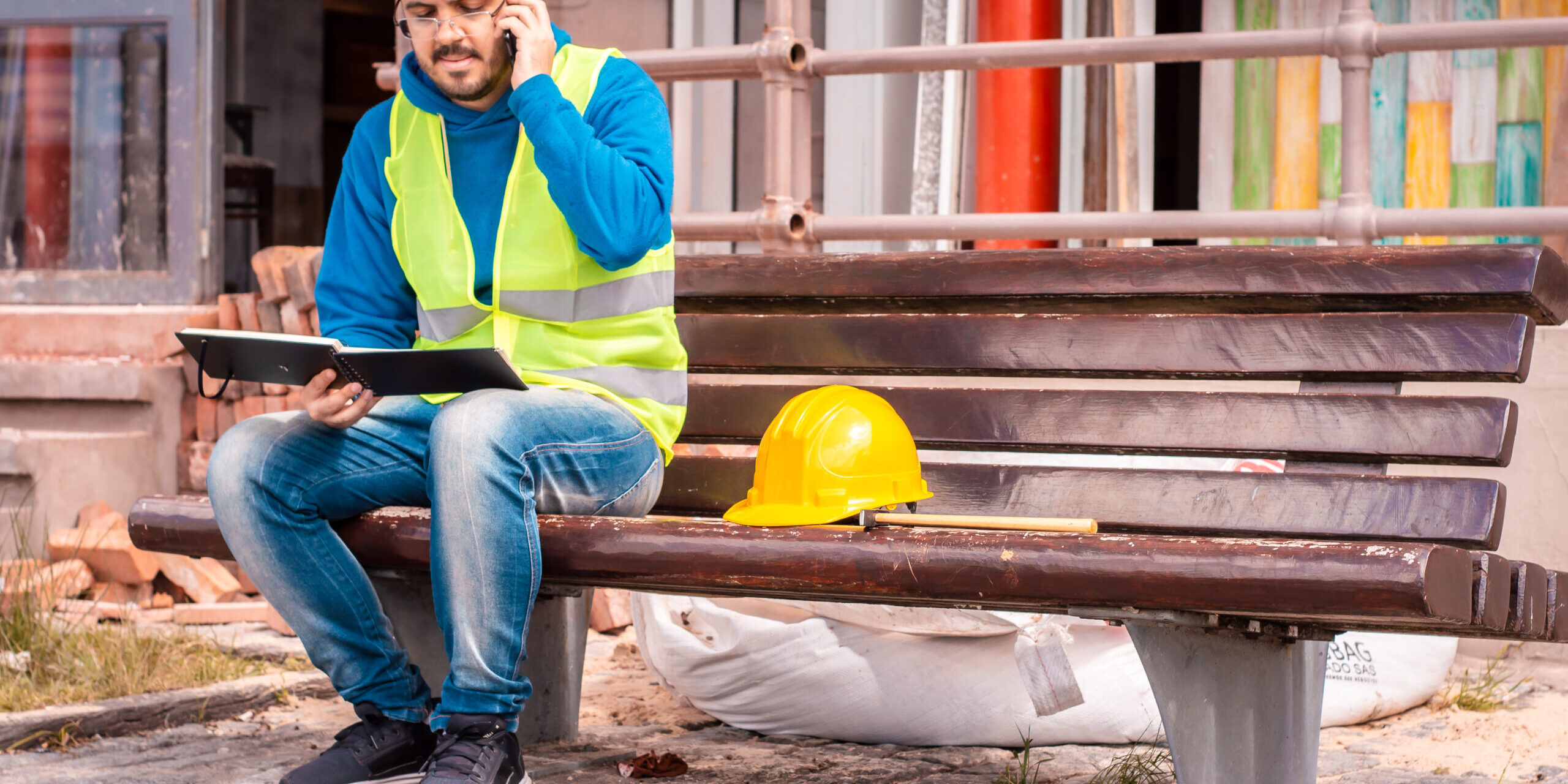 Construction worker in a yellow safety vest sitting on a bench, talking on the phone and reviewing a notebook, with a yellow hard hat beside him