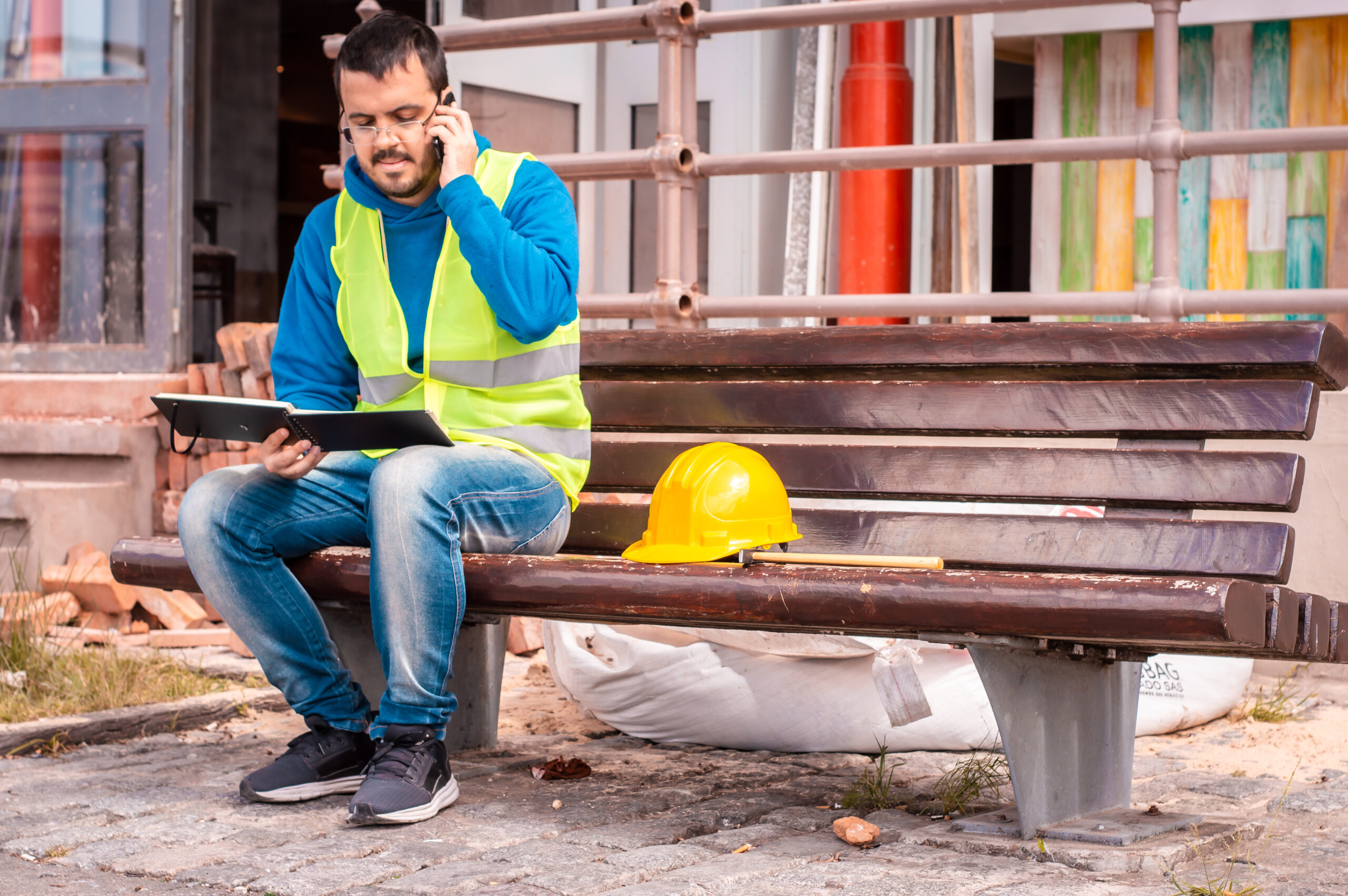 Construction worker in a yellow safety vest sitting on a bench, talking on the phone and reviewing a notebook, with a yellow hard hat beside him