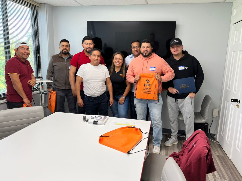 Un grupo de ocho personas en una sala de reuniones posando para una foto. Están frente a una pantalla grande, sosteniendo bolsas y materiales de color naranja con la marca HDS. El grupo está compuesto por hombres y una mujer, todos sonriendo y mirando a la cámara. En primer plano, hay una mesa con artículos de oficina, incluidos unos lentes y una bolsa naranja. El entorno parece ser un taller o seminario de HDS para obtener la licencia de contratista