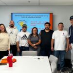 Un grupo de siete personas, incluyendo hombres y una mujer, posa sonriente en un salón de clases frente a una pantalla con un mensaje de felicitaciones por completar el curso de preparación para la licencia de contratista en Maryland. La pantalla tiene fondo azul con texto en rojo y blanco. En la mesa del frente hay un termo rojo, una libreta y bolsas naranjas con el logo de HDS