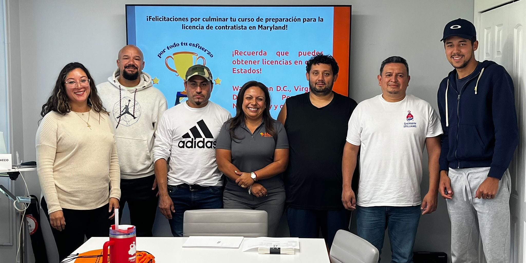 Un grupo de siete personas, incluyendo hombres y una mujer, posa sonriente en un salón de clases frente a una pantalla con un mensaje de felicitaciones por completar el curso de preparación para la licencia de contratista en Maryland. La pantalla tiene fondo azul con texto en rojo y blanco. En la mesa del frente hay un termo rojo, una libreta y bolsas naranjas con el logo de HDS