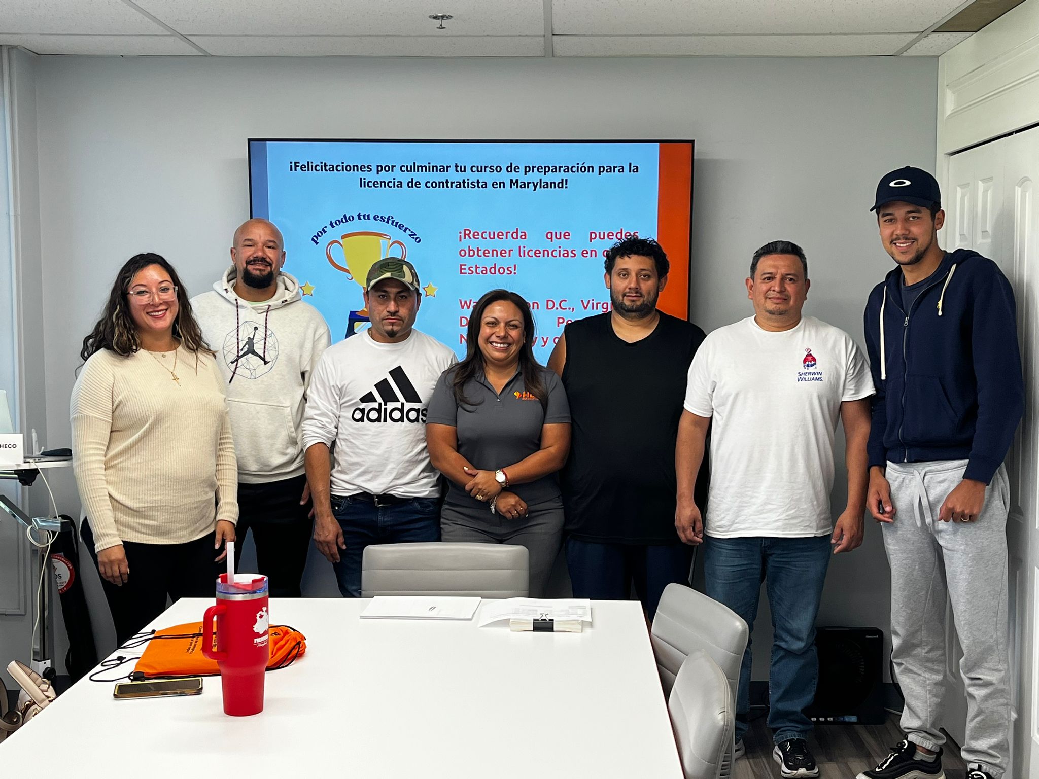 Un grupo de siete personas, incluyendo hombres y una mujer, posa sonriente en un salón de clases frente a una pantalla con un mensaje de felicitaciones por completar el curso de preparación para la licencia de contratista en Maryland. La pantalla tiene fondo azul con texto en rojo y blanco. En la mesa del frente hay un termo rojo, una libreta y bolsas naranjas con el logo de HDS