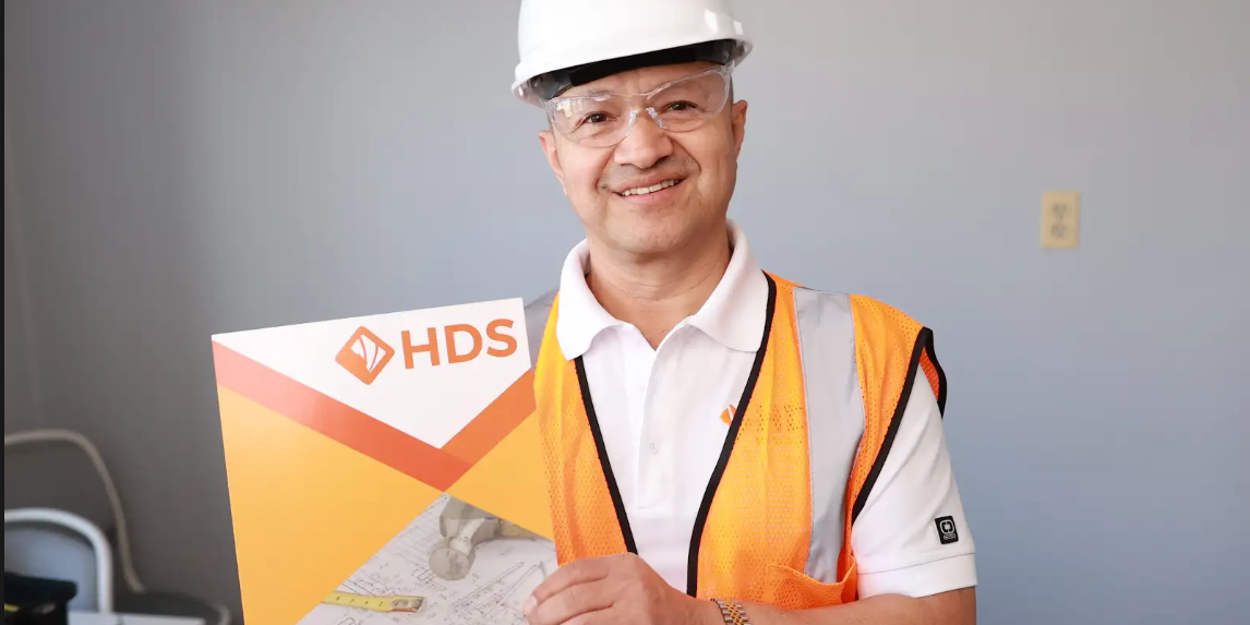 Un hombre sonriente con casco blanco y chaleco de seguridad naranja sostiene una carpeta con el logo de HDS. La carpeta muestra planos de construcción y herramientas, destacando un entorno profesional relacionado con la industria de la construcción, licencia de contratista o contractor license