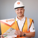 Un hombre sonriente con casco blanco y chaleco de seguridad naranja sostiene una carpeta con el logo de HDS. La carpeta muestra planos de construcción y herramientas, destacando un entorno profesional relacionado con la industria de la construcción, licencia de contratista o contractor license