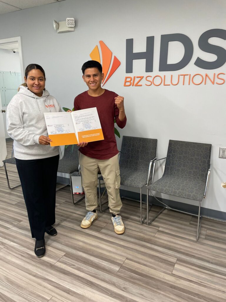 A young man wearing a burgundy shirt and beige pants smiles and proudly holds a folder with documents, raising his fist in a celebratory gesture. Beside him, a woman wearing a white hoodie with the HDS Biz Solutions logo also holds the folder and smiles warmly. They are standing in the modern and clean office of HDS Biz Solutions, with the company’s logo prominently displayed on the wall behind them. The office features gray chairs, a light wood floor, and a potted plant in the corner, reflecting a professional and welcoming environment