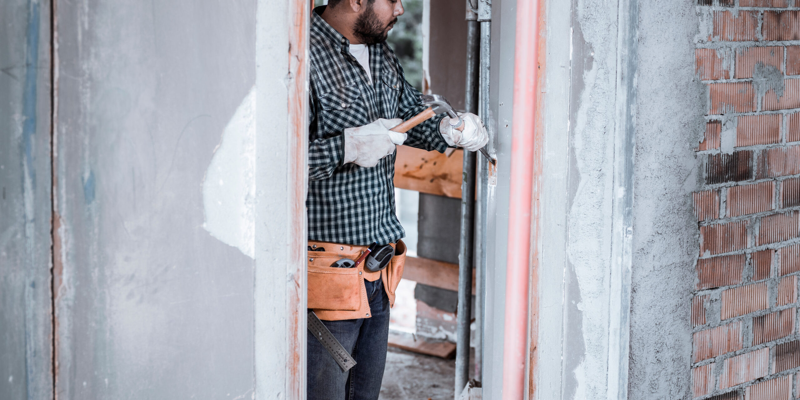 Primer plano de un trabajador de la construcción utilizando un taladro eléctrico sobre una superficie de madera, con virutas de madera en primer plano y un fondo desenfocado que muestra una ventana y luz natural