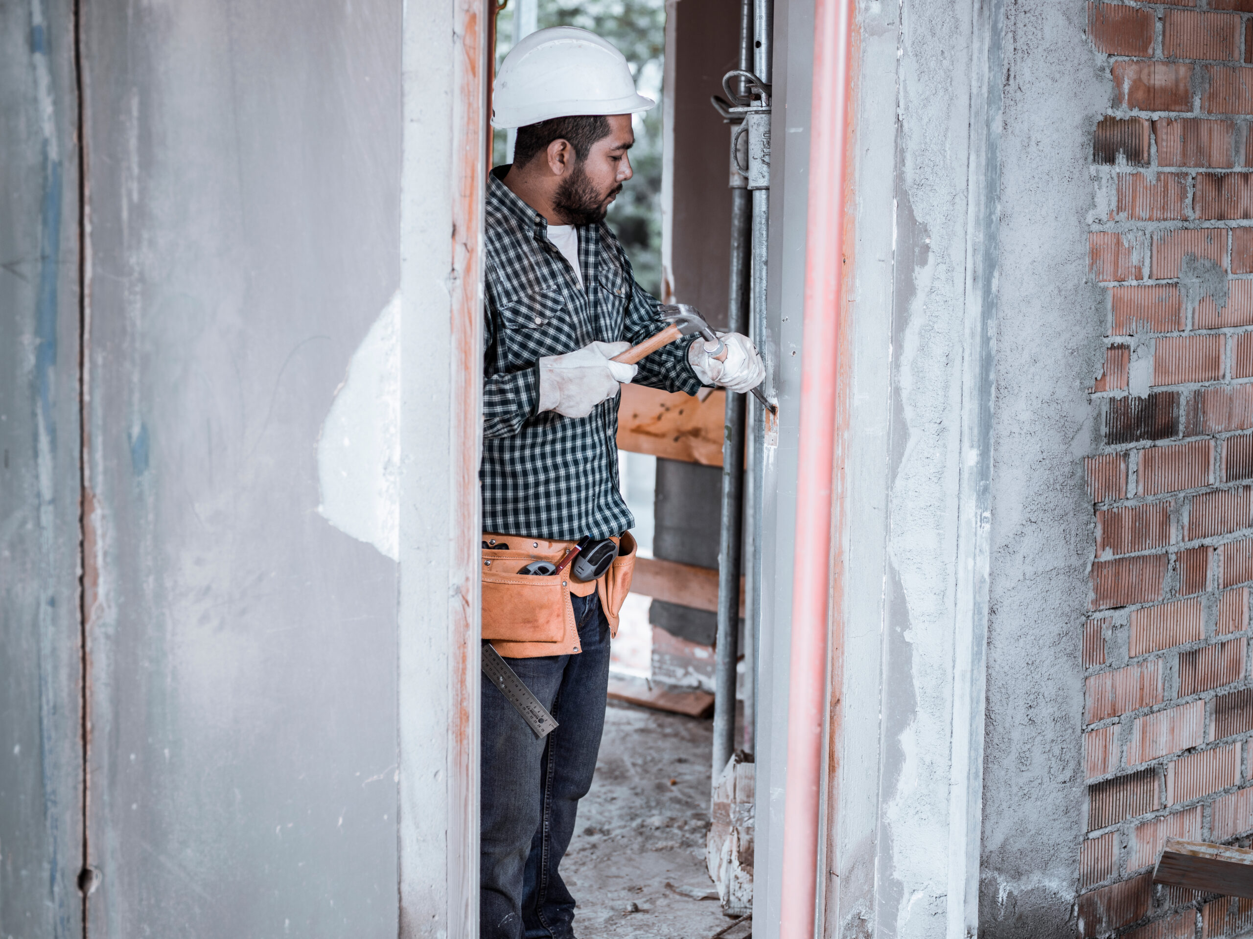 Primer plano de un trabajador de la construcción utilizando un taladro eléctrico sobre una superficie de madera, con virutas de madera en primer plano y un fondo desenfocado que muestra una ventana y luz natural