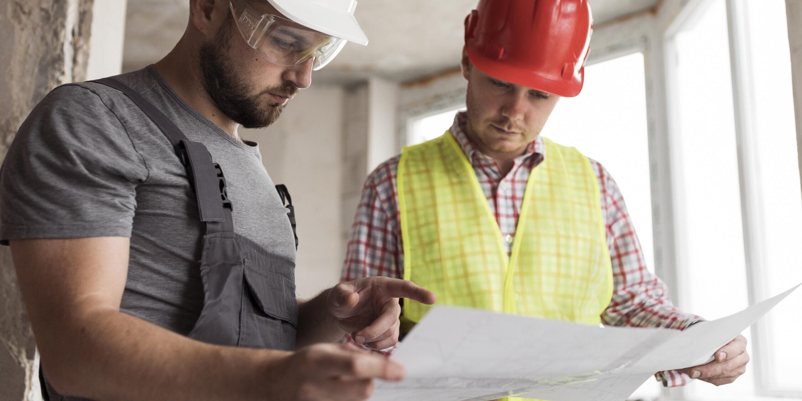contratistas con chalecos reflectantes y cascos de seguridad revisan un proyecto en una tableta en el interior de un edificio en construcción; una de las personas sostiene planos