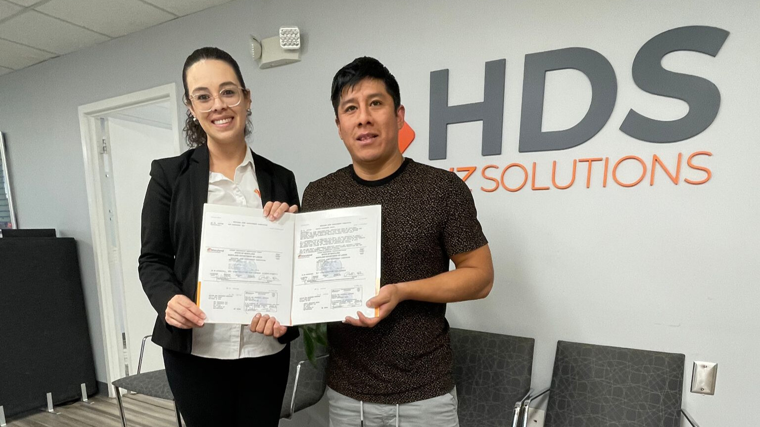 A woman wearing glasses, a white blouse, and a black blazer, and a man in a dark shirt smile while posing in an office at HDS Biz Solutions. Both hold certificates or official documents in front of the company’s gray and orange logo on the wall behind them. The setting is an office with light-colored walls, a paneled ceiling, and chairs in the background.