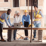 Cuatro personas en un sitio de construcción revisan planos sobre una mesa; dos de ellos llevan chalecos reflectantes, dos usan cascos de seguridad, y una computadora portátil está abierta frente a ellos