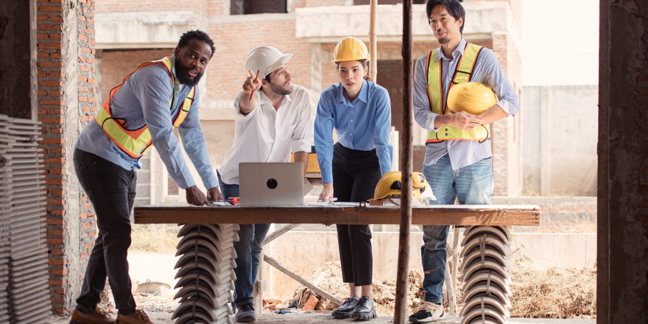 Cuatro personas en un sitio de construcción revisan planos sobre una mesa; dos de ellos llevan chalecos reflectantes, dos usan cascos de seguridad, y una computadora portátil está abierta frente a ellos