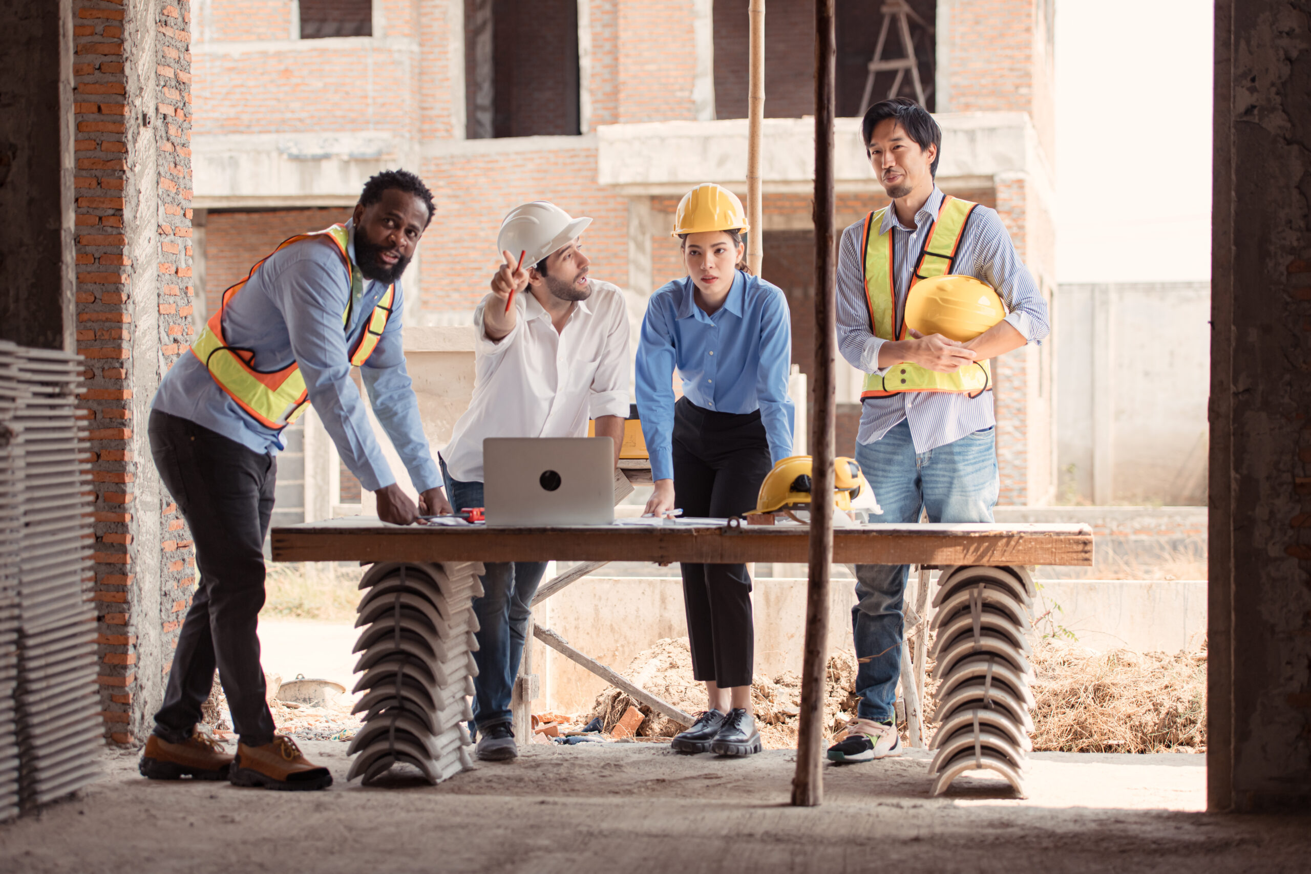 Cuatro personas en un sitio de construcción revisan planos sobre una mesa; dos de ellos llevan chalecos reflectantes, dos usan cascos de seguridad, y una computadora portátil está abierta frente a ellos
